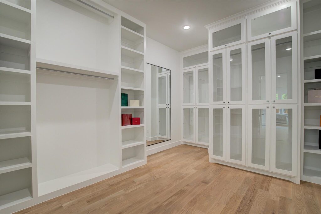 Spacious white walk-in closet with wooden flooring and glass-paneled cabinets.